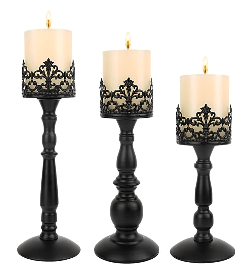 Three elegant matte black iron pillar candle stands of staggered heights, each featuring an intricate, ornate crown-like filigree design holding a lit ivory pillar candle against a white background.