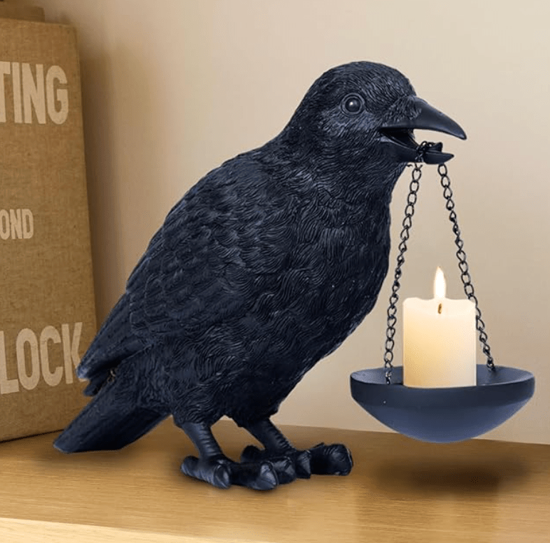 A realistic, textured matte black raven figurine standing on a wooden shelf, holding a black metal chain with a hanging cup and a lit cream-colored candle in its open beak.